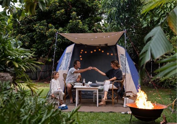 Couple in backyard with daughter enjoying dinner under string lights and covered tent with firepit in foreground